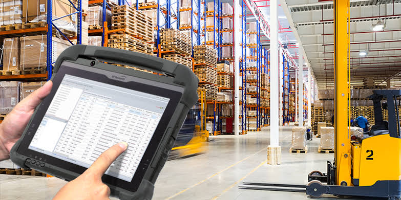 Warehouse worker checking stock on a rugged tablet beside pallet racking and a forklift